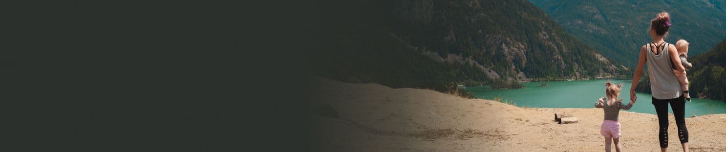 A woman holding a toddler and walking hand-in-hand with a young child, overlooking a scenic lake surrounded by mountains.